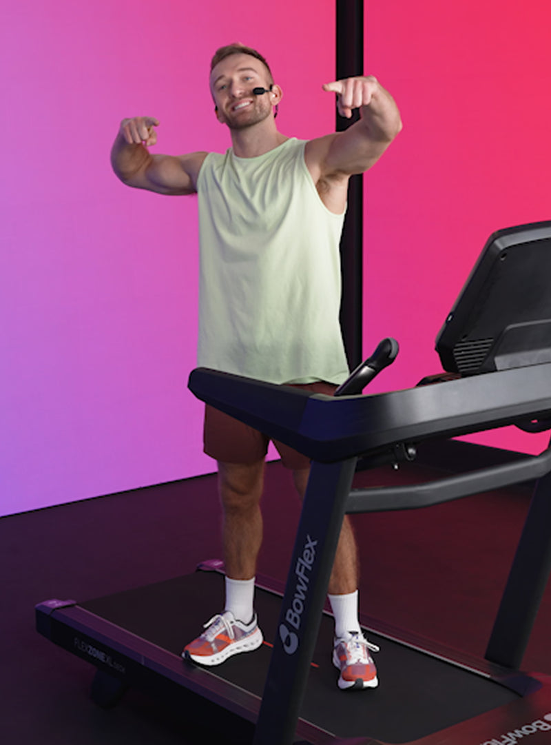 Person standing on a BowFlex treadmill and smiling, with his arms raised and index fingers pointing ahead.
