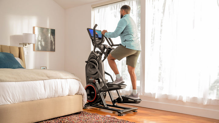 A man is exercising on a BowFlex Max Trainer M6 in a bright room with large windows and a bed.