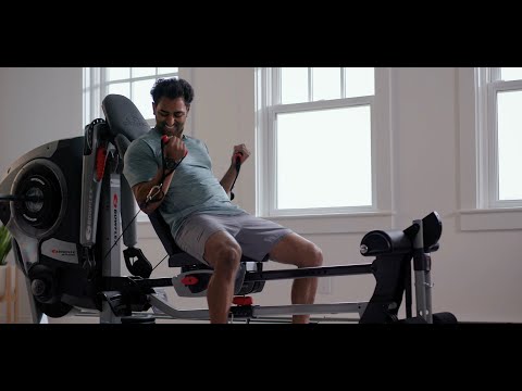 A man performing an arm workout on the BowFlex Revolution Home Gym with its leg press station attached in a bright, modern room with wood floors and large windows.