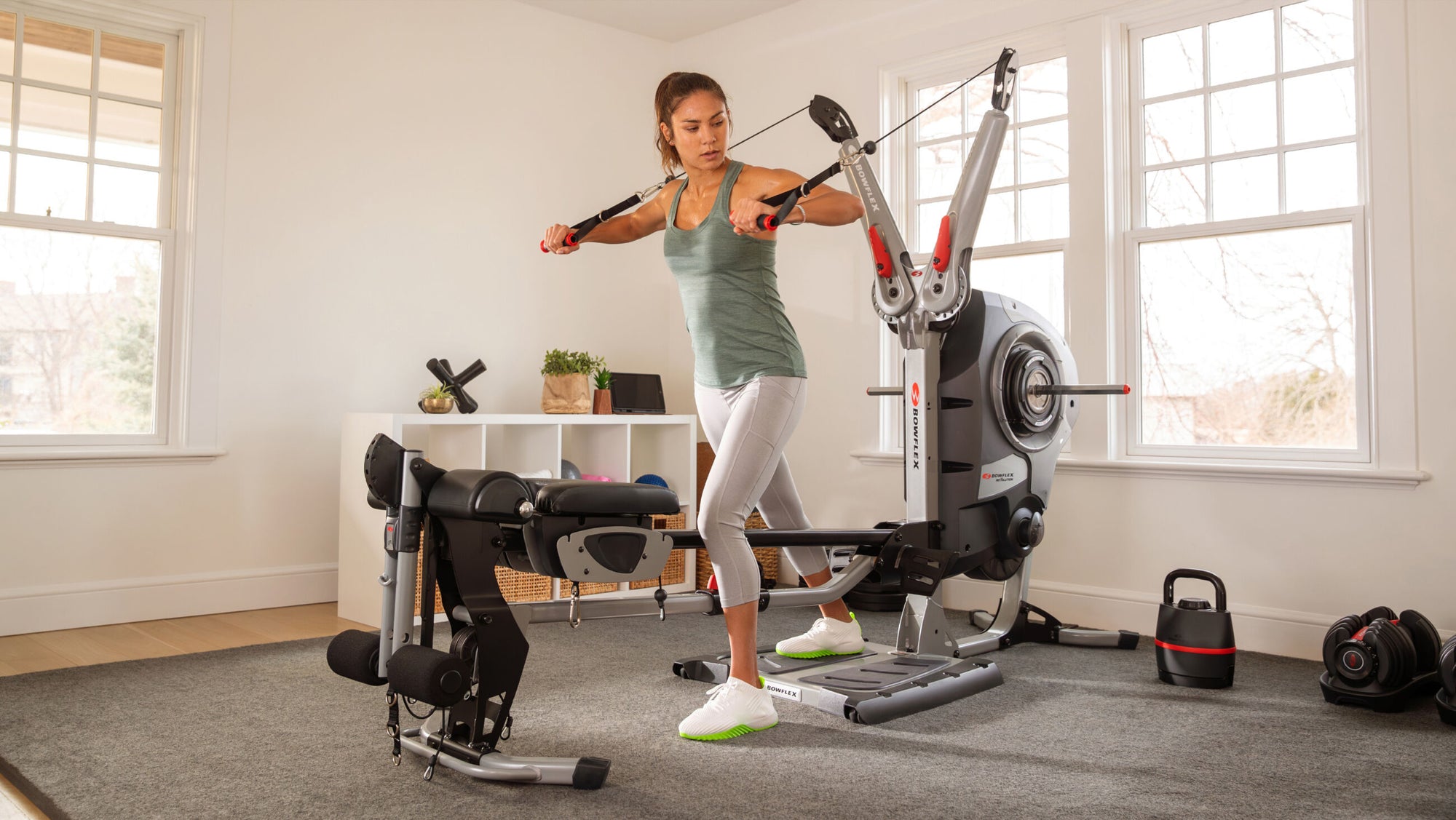 A woman performing an arm workout on the BowFlex Revolution Home Gym with its leg press station attached in a bright, modern room with wood floors and large windows.