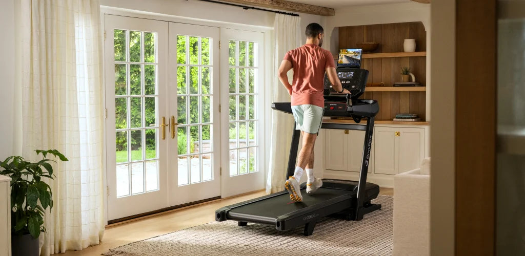 A man running on the BowFlex T9 Treadmill inside a bright, modern room with large white French doors, wooden shelves, and a light-colored rug.
