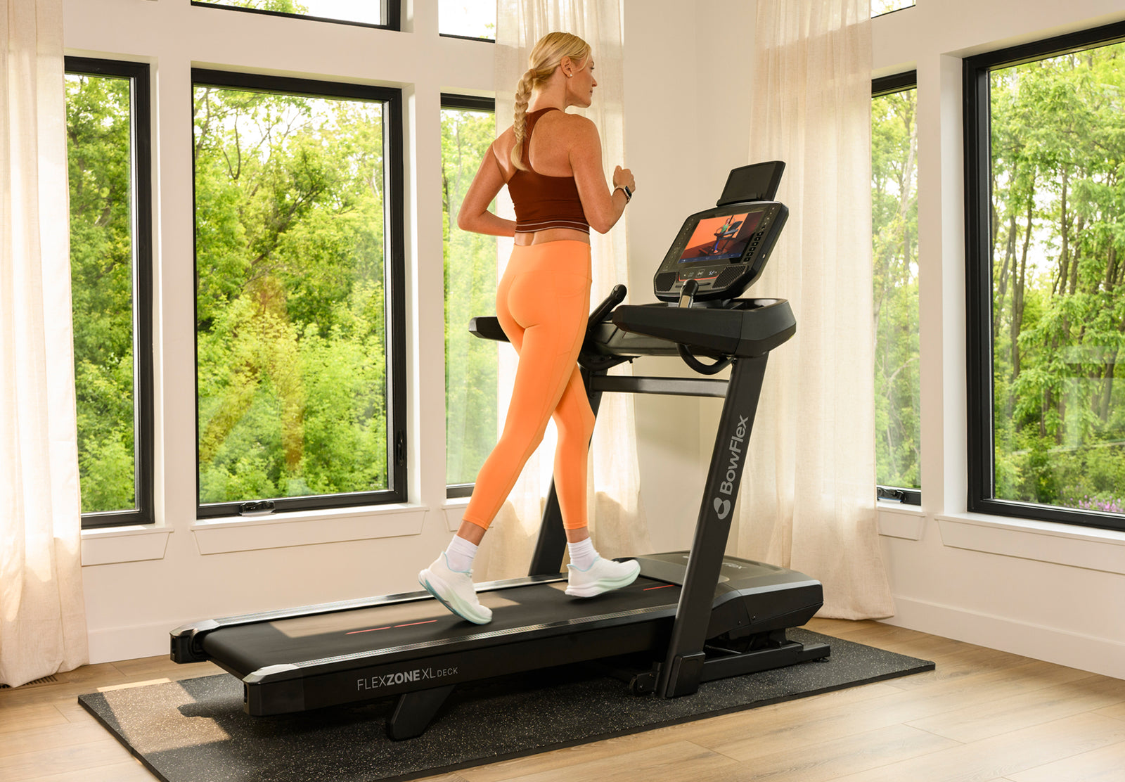 A woman in athletic wear runs on a BowFlex T16 Treadmill in a bright room with large windows and green outdoor views.