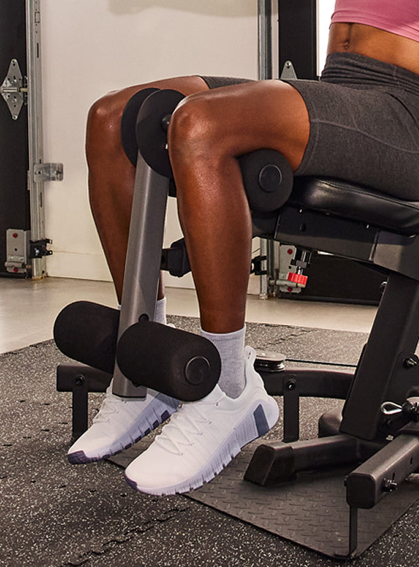 A close-up of a person's legs, wearing white sneakers and gray shorts, resting on the BowFlex Xtreme 2 SE leg extension system in a gym.