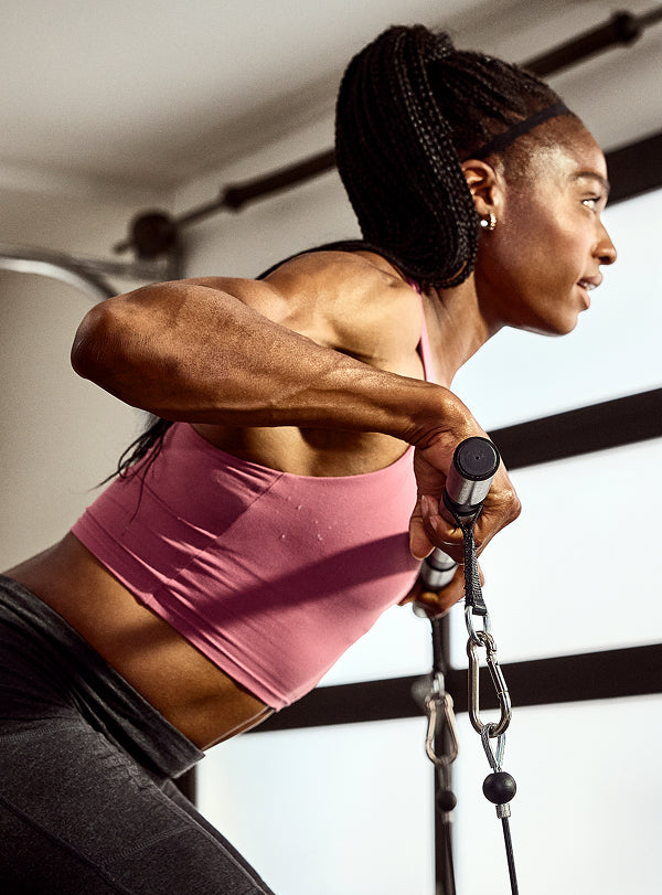 A woman in a pink sports bra and dark gray leggings exercises with the BowFlex Xtreme 2 SE 6-grip steel straight bar in a home gym.