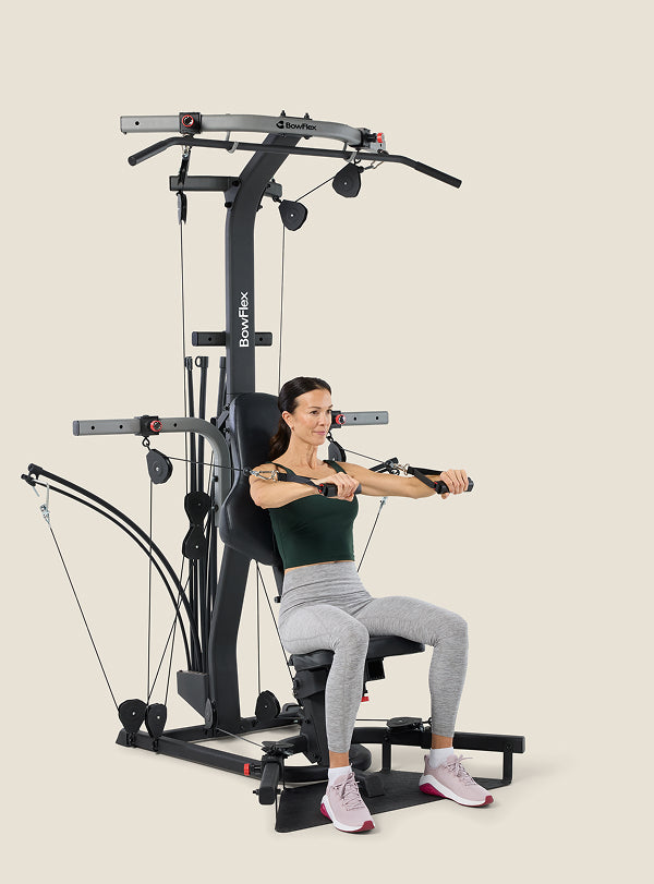 Woman seated on the BowFlex Xtreme 2 SE Home Gym, arms out in front of her while holding onto the cable pulleys.