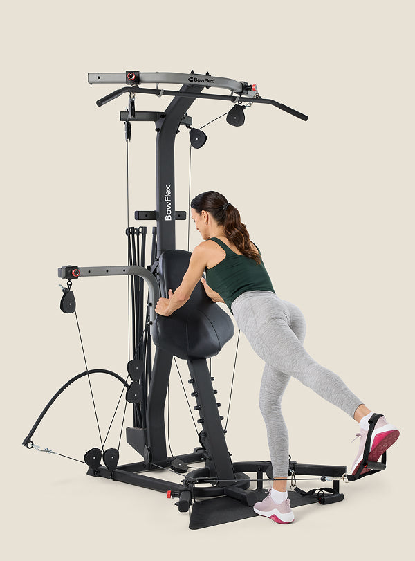 Woman facing and leaning on the BowFlex Xtreme 2 SE Home Gym, using the cable pulley system for a leg workout.