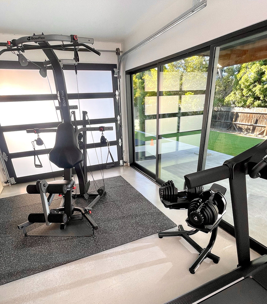 The Bowflex Xtreme 2 SE inside a garage and a weight bench with barbell rack, set on a dark gray gym mat. Large glass sliding doors reveal a backyard with green grass and trees.