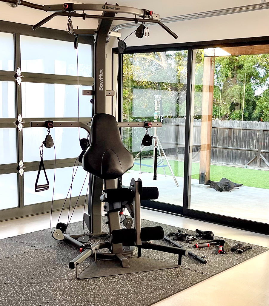 The BowFlex Xtreme 2 SE home gym with a black padded seat and multiple attachment handles, placed on a gray rug in front of large glass sliding doors overlooking a backyard.