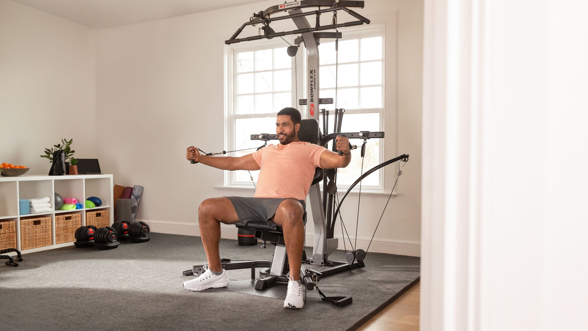 A man wearing a peach t-shirt and gray shorts sits on BowFlex Xtreme 2 SE, pulling its dual purpose grips toward his chest in a brightly lit room with a large window.