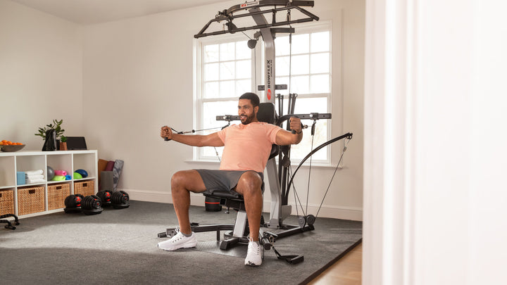 A man wearing a peach t-shirt and gray shorts sits on BowFlex Xtreme 2 SE, pulling its dual purpose grips toward his chest in a brightly lit room with a large window.