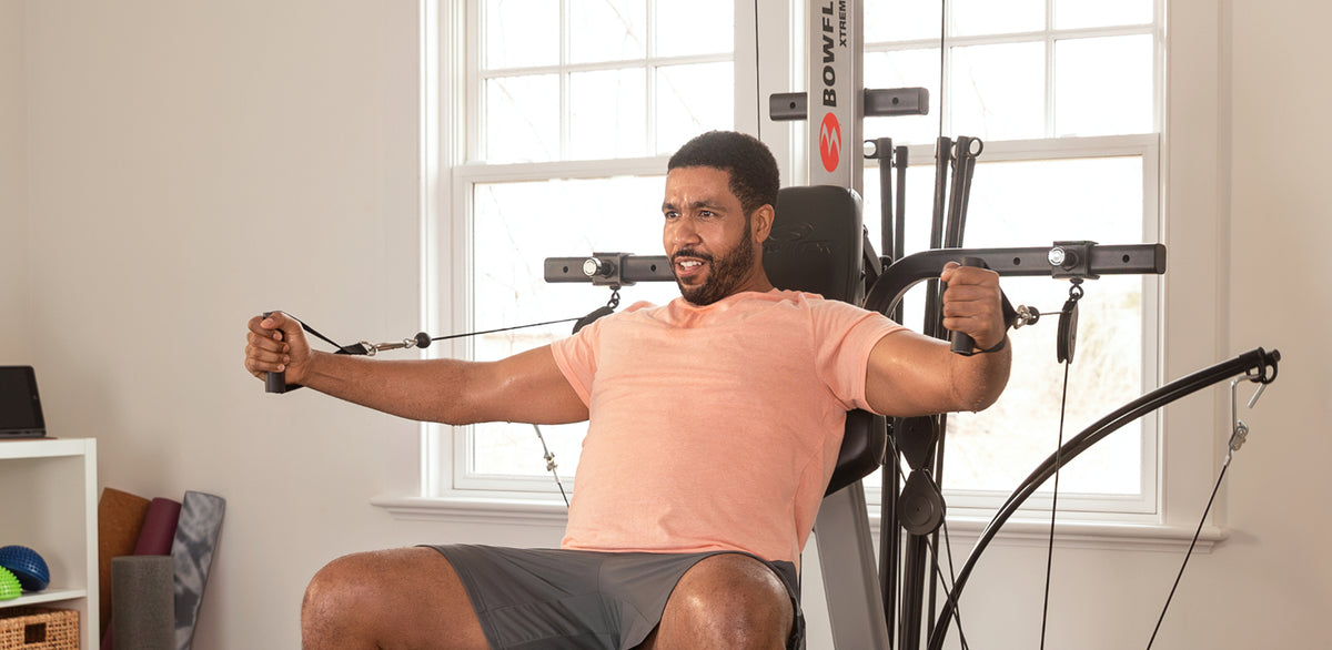 A man wearing a peach t-shirt and gray shorts sits on BowFlex Xtreme 2 SE, pulling its dual purpose grips toward his chest in a brightly lit room with a large window.
